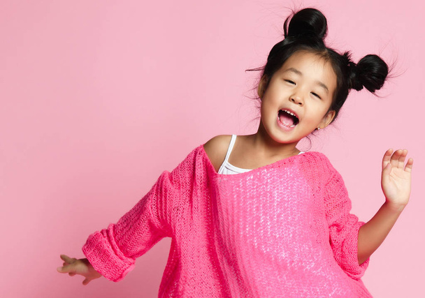 Asian kid girl in pink sweater, white pants and funny buns sings. close up. - Foto, Imagen