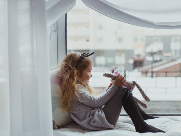 Pretty girl sitting near window and playing with soft toy, intdoor - Fotoğraf, Görsel