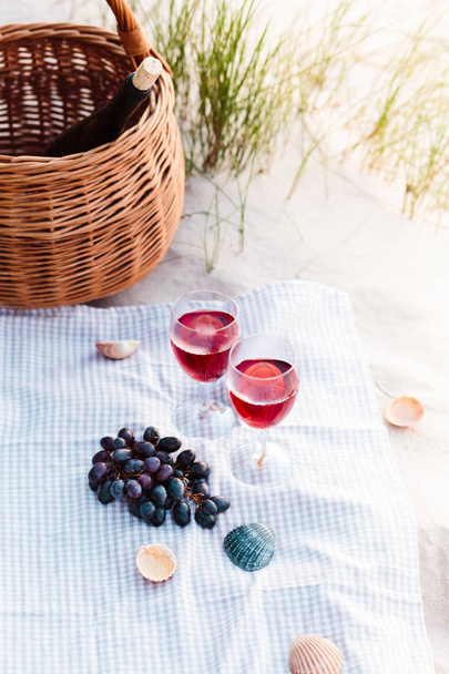 Two wine glasses with red wine standing on cloth, on beach, beside grapes and wicker basket with bottle of wine - Valokuva, kuva