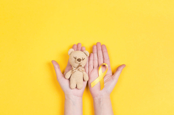 Girl hands holding children's toy with a Childhood Cancer Awareness Yellow Ribbon on yellow background. Childhood Cancer Day February, 15. - Photo, image