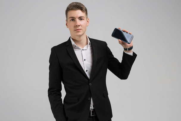 Portret van de jonge mens in zwart pak toont mobiele telefoon met leeg scherm geïsoleerd op witte achtergrond - Foto, afbeelding
