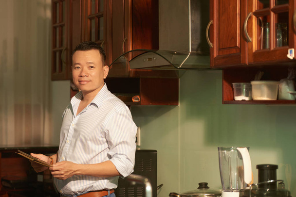 デジタル タブレットの彼の手の中で台所に立っている陽気な成熟したアジアの男 - 写真・画像