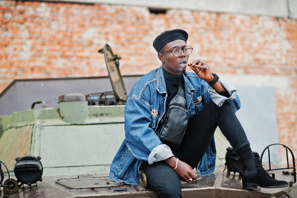 African american man in jeans jacket, beret and eyeglasses, smoking cigar and posed against btr military armored vehicle. - Foto, Imagem