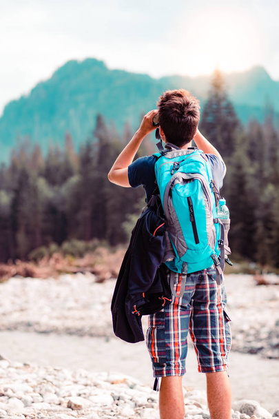 Young wanderer with backpack looks through a binoculars on mountains peaks, stands over a river. Boy spends a vacation in mountains, wandering with backpack, he is wearing sports summer clothes - Fotografie, Obrázek