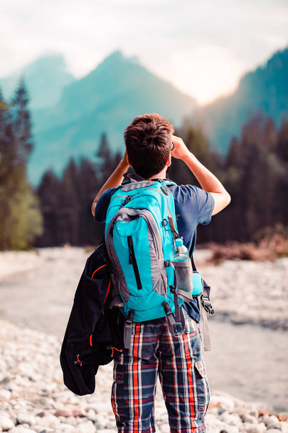Young wanderer with backpack looks through a binoculars on mountains peaks, stands over a river. Boy spends a vacation in mountains, wandering with backpack, he is wearing sports summer clothes - Foto, Bild