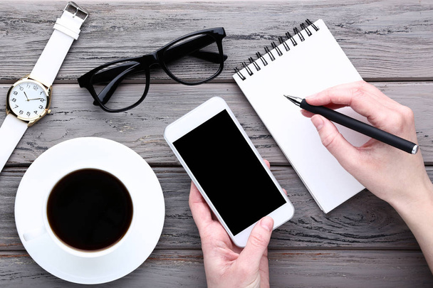 Smartphone in female hand with cup of coffee, notebook and wrist watch on wooden table - Photo, image