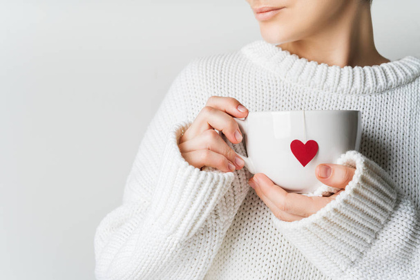 Close view of woman in warm knitted woolen sweater holding white mug in hands with heart of tea bag with love, Valentine's day mockup - Фото, изображение