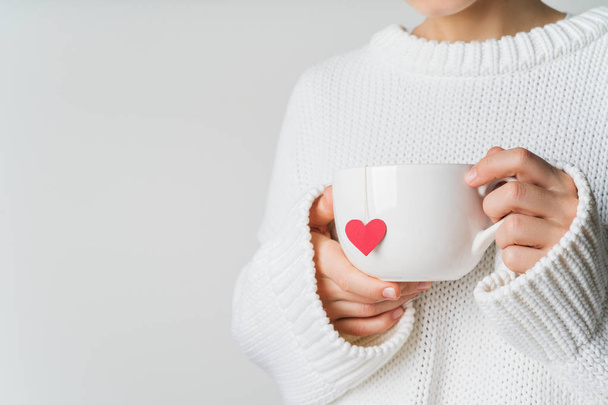 Close view of woman in warm knitted woolen sweater holding white mug in hands with heart of tea bag with love, Valentine's day mockup - Фото, зображення