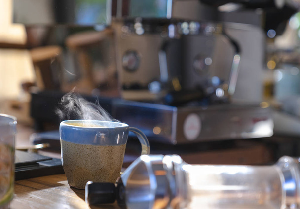 close up a ceramic cup of hot smoke coffee on wooden table with blurred coffee machine in background - Photo, Image