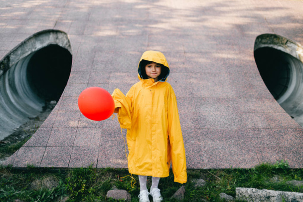 little girl in oversized yellow raincoat with red balloon against sewage tunnel pipe - Foto, Imagem