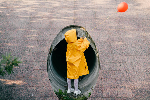 little girl in oversized yellow raincoat with red balloon against sewage tunnel pipe - 写真・画像