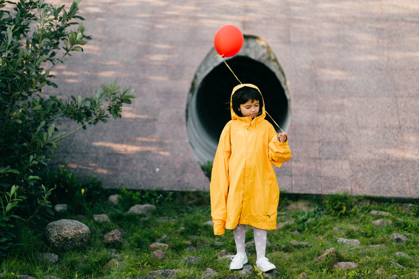 little girl in oversized yellow raincoat with red balloon against sewage tunnel pipe - Valokuva, kuva