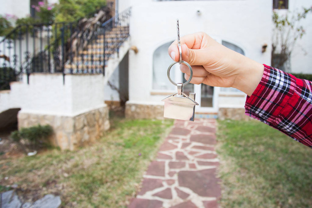Ακίνητη περιουσία και το κατάλυμα έννοια - χέρι κρατά κλειδιά σπιτιών στο σπίτι σε σχήμα μπρελόκ μπροστά σε ένα νέο σπίτι - Φωτογραφία, εικόνα