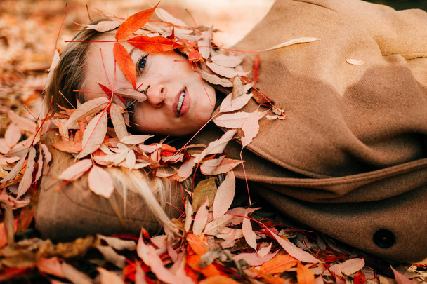 portrait of beautiful young woman in woollen coat lying on ground with fallen leaves around - Foto, Imagem