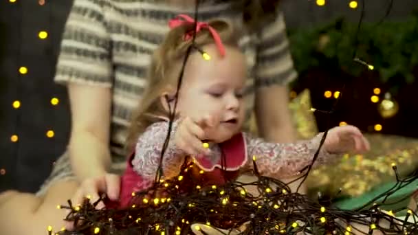 Petite fille bébé et sa mère assis sur un fond d'arbre de Noël décoré et jouant avec des guirlandes de Noël. Bel enfant jouant avec des guirlandes avec des lumières jaunes près de l'épinette verte
. - Séquence, vidéo