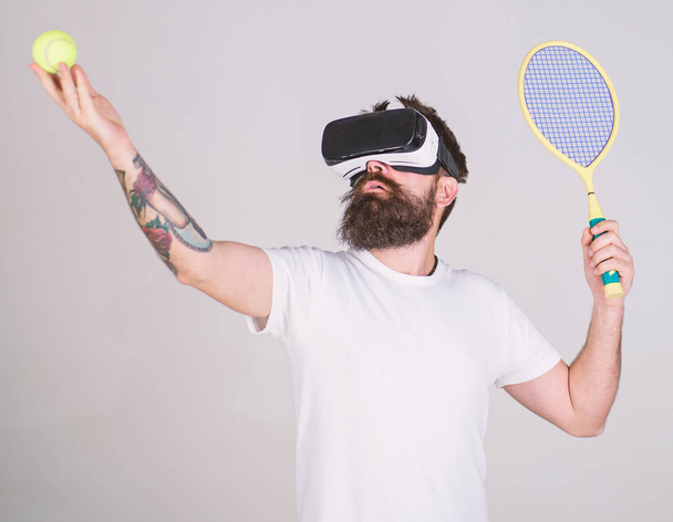 Man with beard in VR glasses, grey background. Hipster on concentrated face use modern technology for sport games. Virtual tennis concept. Guy with VR glasses play tennis with racket and ball - Fotografie, Obrázek