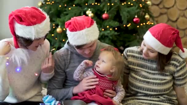 Close friends sitting on the carpet with a baby girll, playing with her and garland near the Christmas tree, dressed in Santa hat. Three adults and one child near decorated spruce. - Filmagem, Vídeo