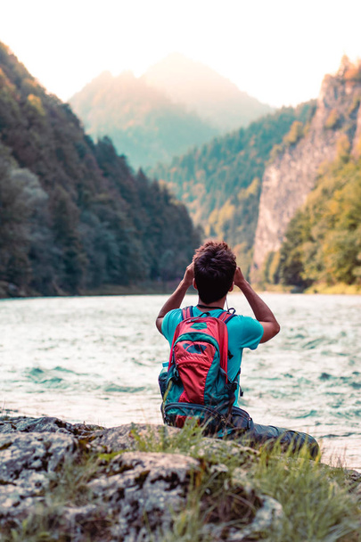 Ein junger Tourist mit Rucksack blickt durch ein Fernglas auf Berggipfel, steht auf einem Felsen über einem Fluss. Junge verbringt Urlaub in den Bergen, wandert mit Rucksack, trägt Sommerkleidung - Foto, Bild