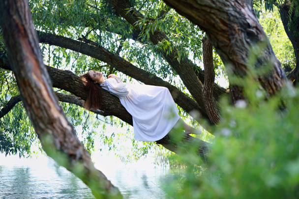 Beautiful woman in white dress lying on a tree over the river - Photo, Image