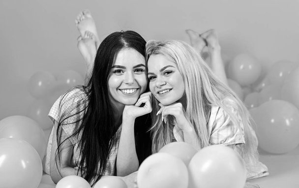 Friendship concept. Blonde and brunette on smiling faces have fun at domestic party. Girls lay on belly near balloons, pink background. Sisters or best friends in pajamas at girlish pajamas party - Φωτογραφία, εικόνα
