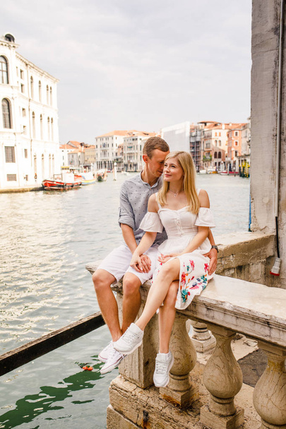 Young beautiful couple, girl in white dress and man in blue shirt are sitting on the balcony in the beautiful city of Venice in Italy - Foto, afbeelding