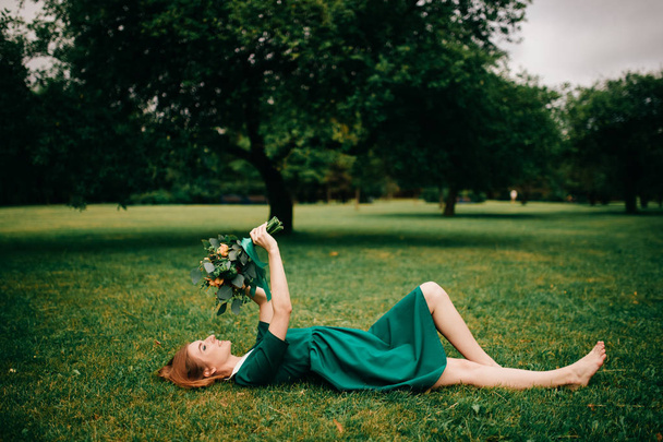 young red hair woman in green dress with bouquet of summer flowers lying on green grass - Photo, Image