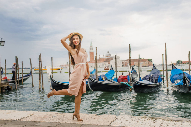 Menina bonita nova com um vestido rosa está de pé perto da água com vista para as gôndolas e bela Veneza
 - Foto, Imagem