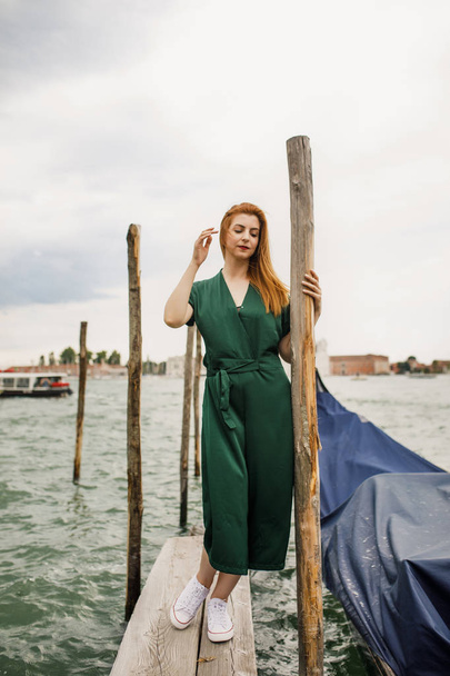 Mooi meisje met rode haren in groene kleren staat in de buurt van het water met uitzicht op de prachtige Venetië - Foto, afbeelding