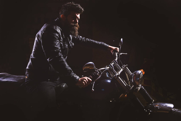 Man with beard, biker in leather jacket sitting on motor bike in darkness, black background. Night racer concept. Macho, brutal biker in leather jacket riding motorcycle at night time, copy space - Fotó, kép