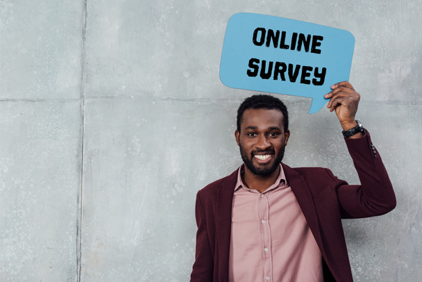 lächelnder afrikanisch-amerikanischer Geschäftsmann, der in die Kamera blickt und eine Sprechblase mit "Online-Umfrage" -Schriftzug auf grauem Hintergrund hält - Foto, Bild