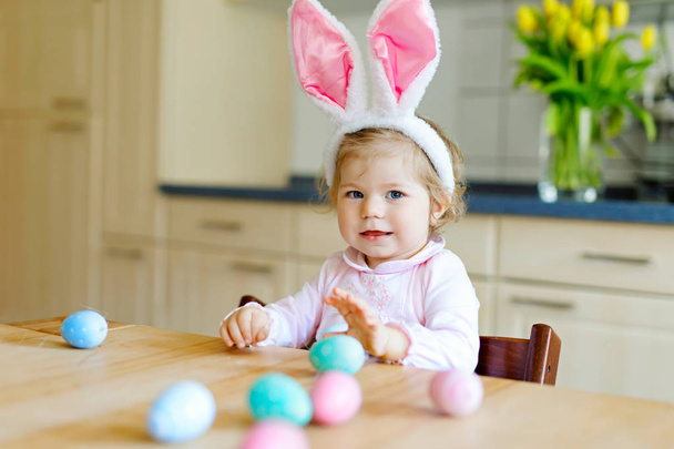 Ładny mały malucha dziewczyna ubrana w Easter bunny uszy gry z kolorowych jaj pastelowych. Szczęśliwe dziecko dziecko rozpakowywania prezentów. Adorable kid w różowe ubrania wakacje - Zdjęcie, obraz