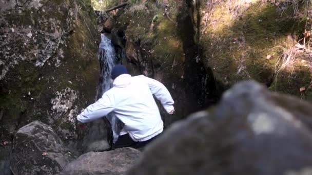 Widok mężczyzny w białą kurtkę, idąc w dół do wodospadu w wąwozie z góry z tyłu. Turystycznych, stojąc na kamienie i patrzy na przepływ wody spadające w wąwozie. - Materiał filmowy, wideo