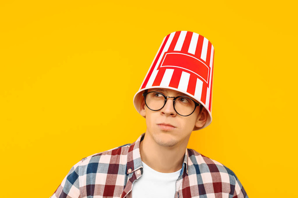 Cheerful man in glasses is happy to watch a movie, with a bucket of popcorn on his head, on a yellow background - 写真・画像