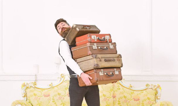 Man with beard and mustache wearing classic suit delivers luggage, luxury white interior background. Butler and service concept. Macho elegant on surprised face carries pile of vintage suitcases - Fotó, kép