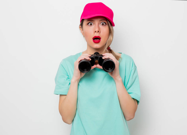 Beautiful young girl in pink cap and blue t-shirt looking for something with binocular on white background. - Photo, Image