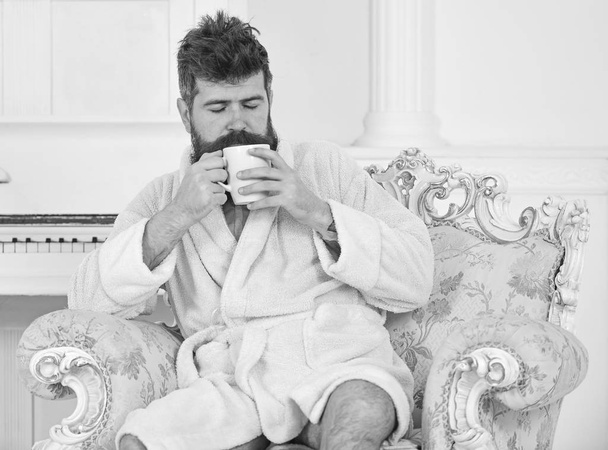Man sleepy in bathrobe, drinks coffee, enjoying aroma in luxury hotel in morning, white background. Man with beard and mustache enjoys morning while sitting on luxury armchair. Elite leisure concept - Foto, Imagen