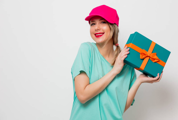 Beautiful young girl in pink cap and blue t-shirt with holiday present box on white background. - Photo, Image
