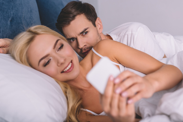 ύποπτες νεαρός peeking χαμογελαστή γυναίκα χρησιμοποιώντας το smartphone στο κρεβάτι, δυσπιστία έννοια  - Φωτογραφία, εικόνα