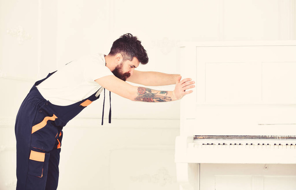 Man with beard and mustache, worker in overalls pushes piano, white background. Delivery service concept. Loader moves piano instrument. Courier delivers furniture in case of move out, relocation - Photo, image