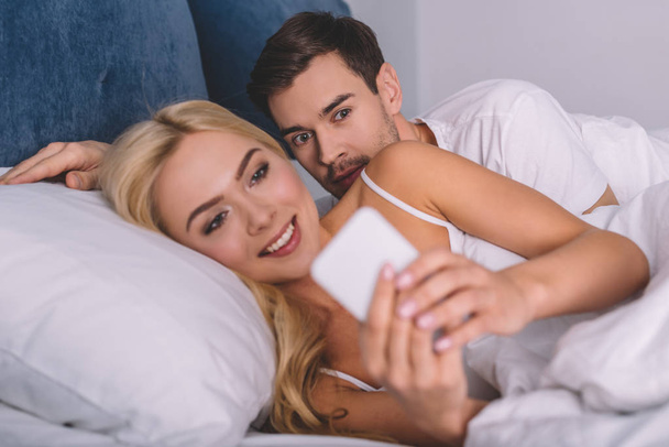 young man peeking at smiling girlfriend using smartphone in bed, distrust concept  - Fotografie, Obrázek