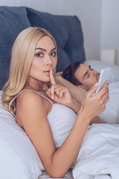 young woman gesturing for silence and looking at camera while boyfriend sleeping in bed, secret concept  - Photo, Image
