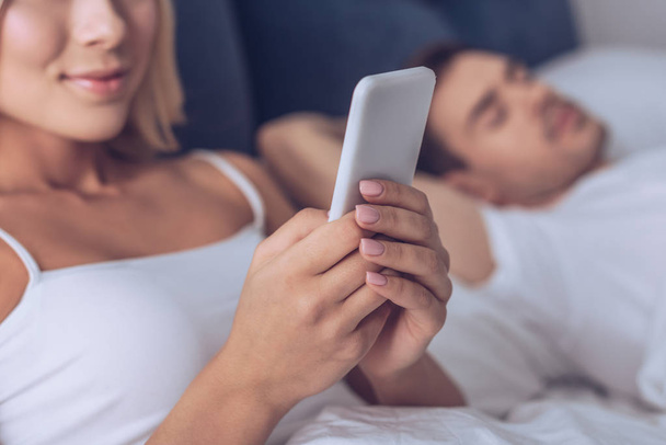 cropped shot of smiling young woman using smartphone while husband sleeping in bed - Photo, Image