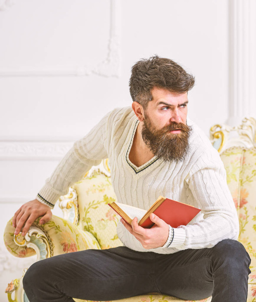Man with beard and mustache sits on baroque style sofa, holds book, white wall background. Reflections on literature concept. Macho on strict face finished reading book. Guy thinking about literature - Photo, image