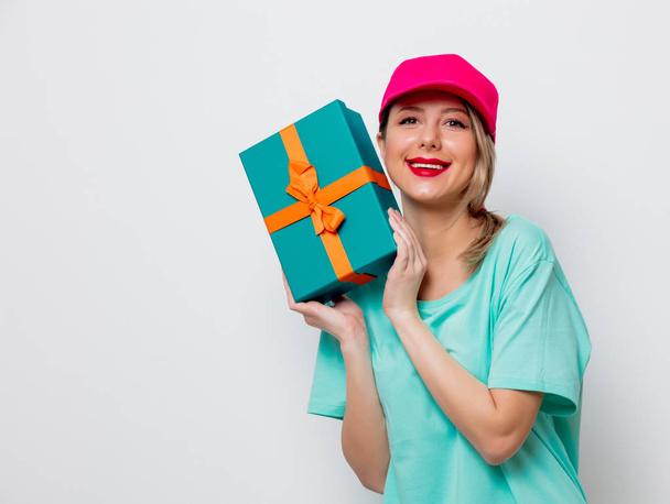 Beautiful young girl in pink cap and blue t-shirt with holiday present box on white background. - Photo, Image