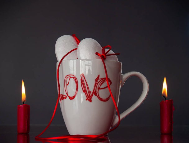Concept of love Two poliespan hearts joined with a red thread that symbolizes the legend of the red thread on a breakfast cup and two red lit candles - Photo, image