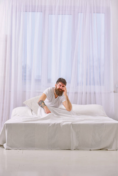 Hangover concept. Guy on disappointed painful face waking up in morning. Man in shirt sits on bed awake, white curtain on background. Macho with beard and mustache suffers from hangover - Fotografie, Obrázek