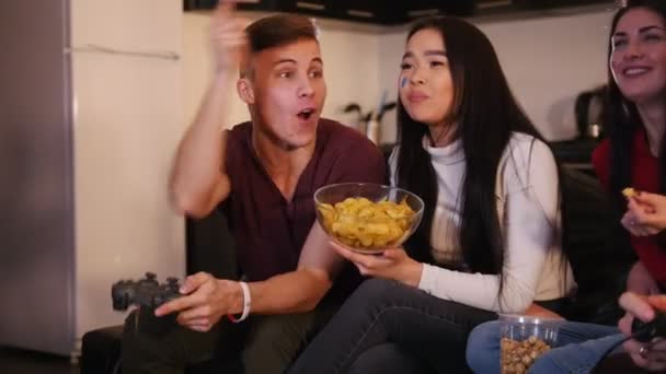 Company of young friends spending time together. Young man enthusiastically playing game holding a joystick. Young woman watching and eating chips - Materiał filmowy, wideo