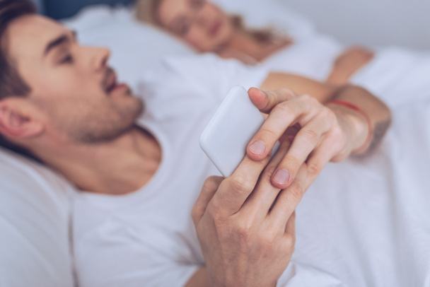 close-up view of young man using smartphone while lying with sleeping wife in bed - Foto, imagen