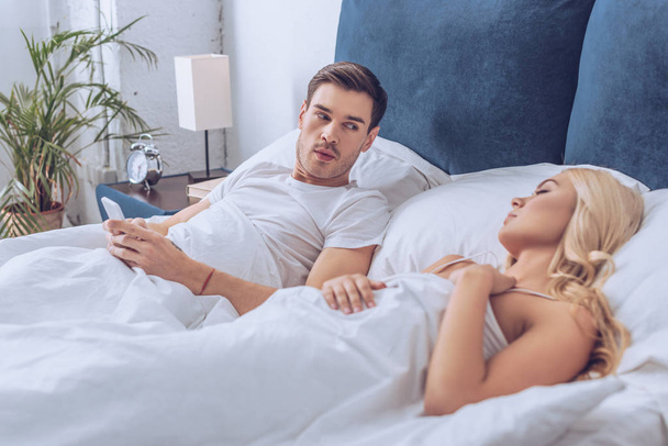 suspicious man lying in bed with smartphone and looking at sleeping girlfriend, mistrust concept  - Photo, Image