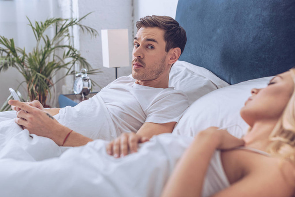 homme en utilisant un smartphone et en regardant la caméra tout en étant couché avec femme endormie au lit
 - Photo, image